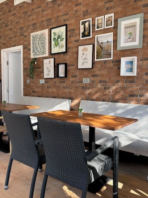 Una habitación con pared de ladrillo decorada con reproducciones botánicas enmarcadas, una mesa de madera y asientos tapizados en gris.