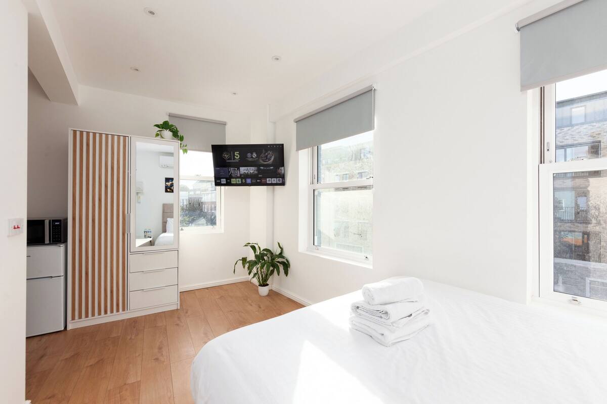 A modern hotel room with a bed, a TV mounted on the wall, a potted plant, and a view of the city through large windows.