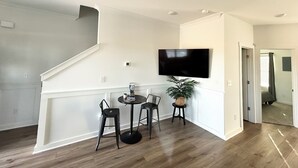 Un salon moderne avec un téléviseur à écran plat fixé au mur, un coin repas comprenant une petite table ronde et deux chaises, et un escalier menant à un autre niveau.