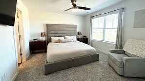 Une chambre avec un grand lit, un ventilateur de plafond, une fenêtre avec des stores, une chaise et une table de chevet avec des lampes.