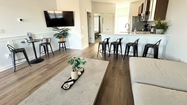 Un salon moderne avec un canapé, une table basse et un téléviseur. On y trouve trois tabourets de bar et une petite table avec une plante. On aperçoit la cuisine au fond, avec un réfrigérateur et un évier.