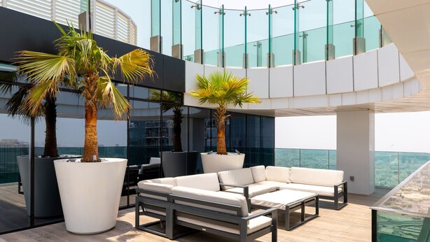 Eine moderne Lounge-Bereich im Freien mit einer weißen Sofagarnitur, zwei großen Kübelpalmen und einem Glasgeländer mit Blick auf eine Stadtlandschaft.