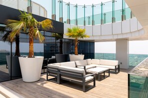 Un espace de détente extérieur moderne, avec un ensemble de canapés blancs, deux grands palmiers en pot et une balustrade en verre donnant sur un paysage urbain.