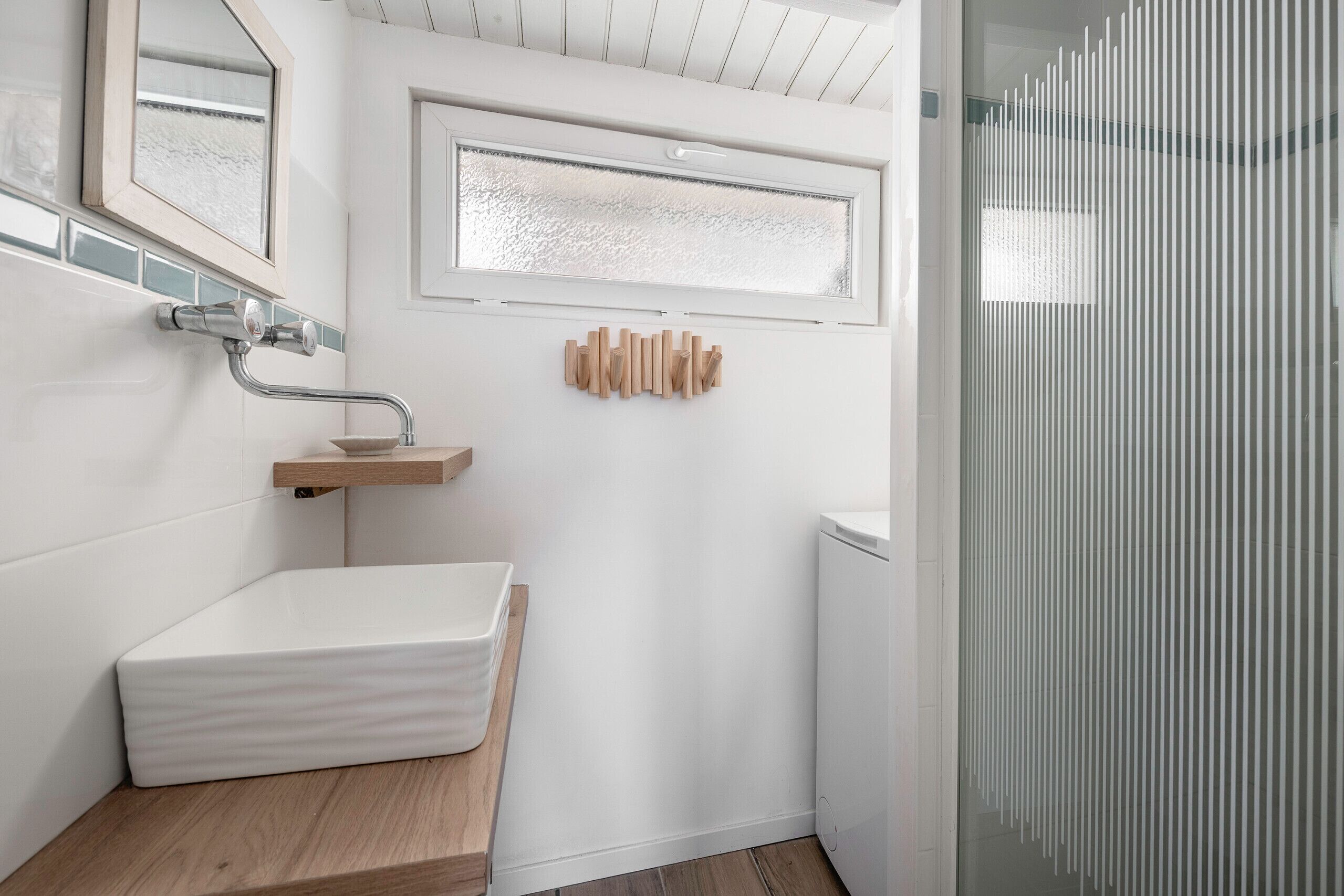 A modern bathroom with a white sink, wooden shelf, and a window above the mirror.