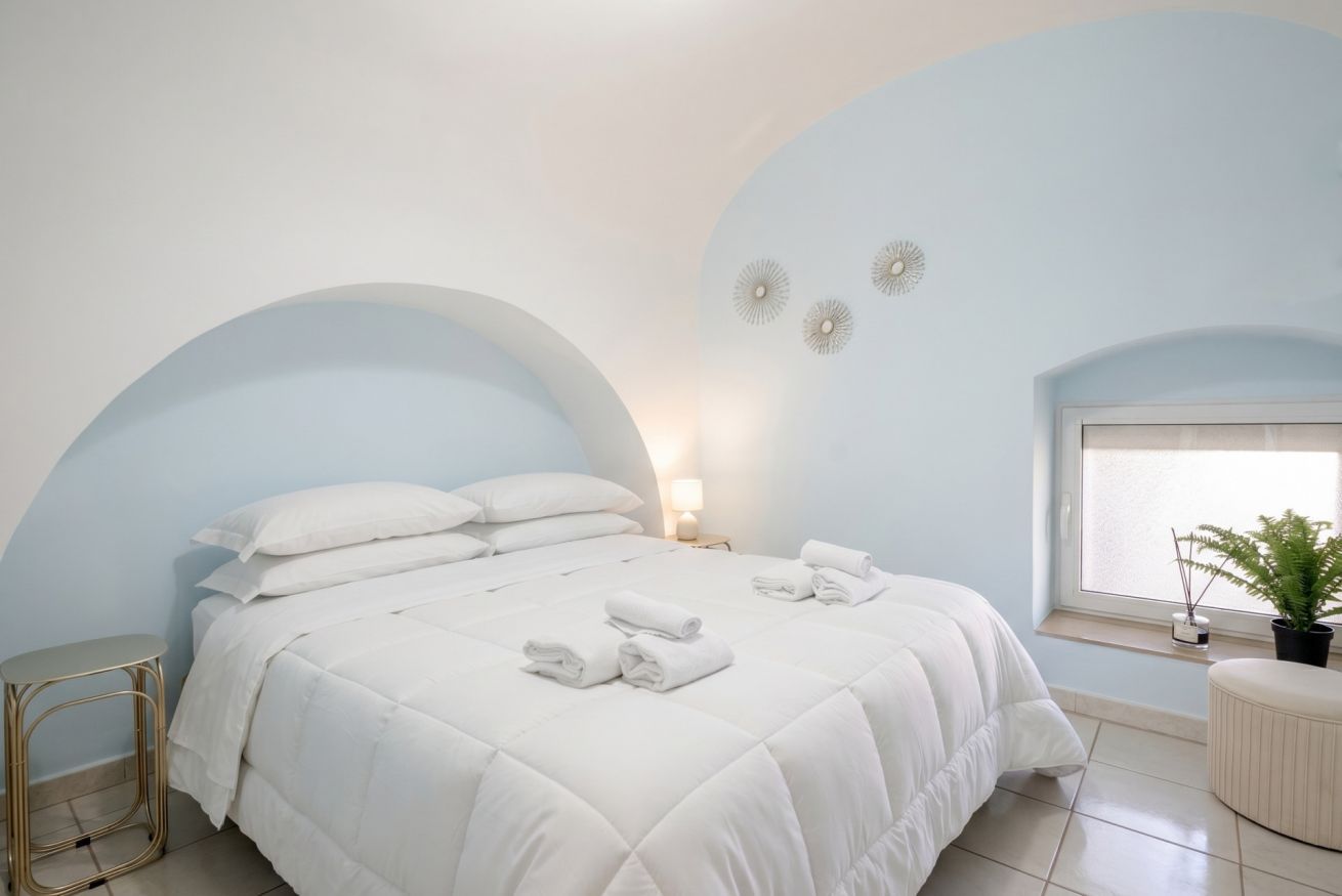 Een slaapkamer met een bed, kussens, een nachtkastje, een lamp, een raam met uitzicht en een plant.