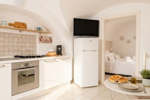 Een kleine keuken met een fornuis, oven en een koffieapparaat, een witte koelkast, en een bed in de aangrenzende kamer.