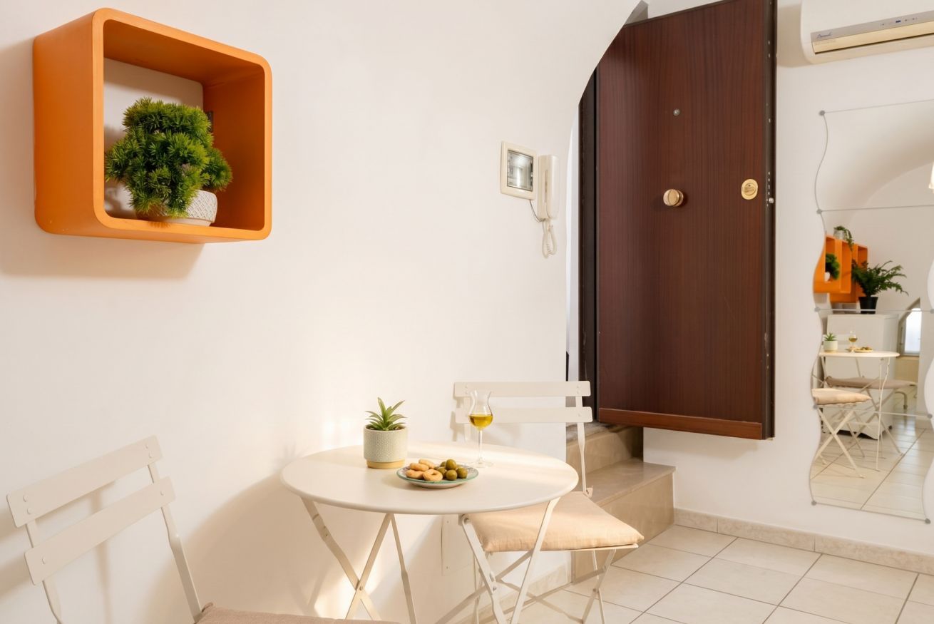 Ein Zimmer mit einem weißen runden Tisch, einer Topfpflanze, einer kleinen Snackplatte, einem weißen Stuhl, einer Holztür und einem weißen an der Wand befestigten Telefon.