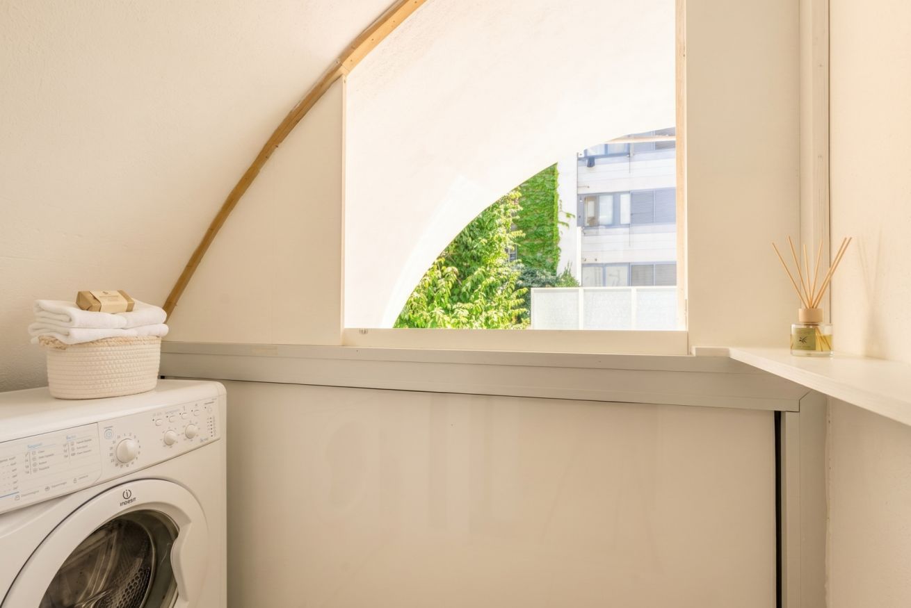 A laundry area with a washing machine, a basket of folded clothes, and a window with a view of greenery and a building.