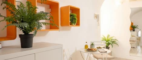 A modern kitchen with white cabinets, a dining area with a table and chairs, and orange shelving units with plants.