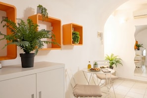 A modern kitchen with white cabinets, a dining area with a table and chairs, and orange shelving units with plants.