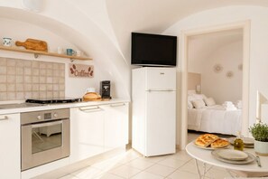 Une cuisine compacte équipée d’une cuisinière, d’un four et d’une machine à café, d’un réfrigérateur blanc, et d’un lit dans la pièce adjacente.