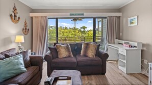 Ein Wohnzimmer mit einem braunen Sofa, einem weißen Schreibtisch und Blick auf Grün durch große Fenster.
