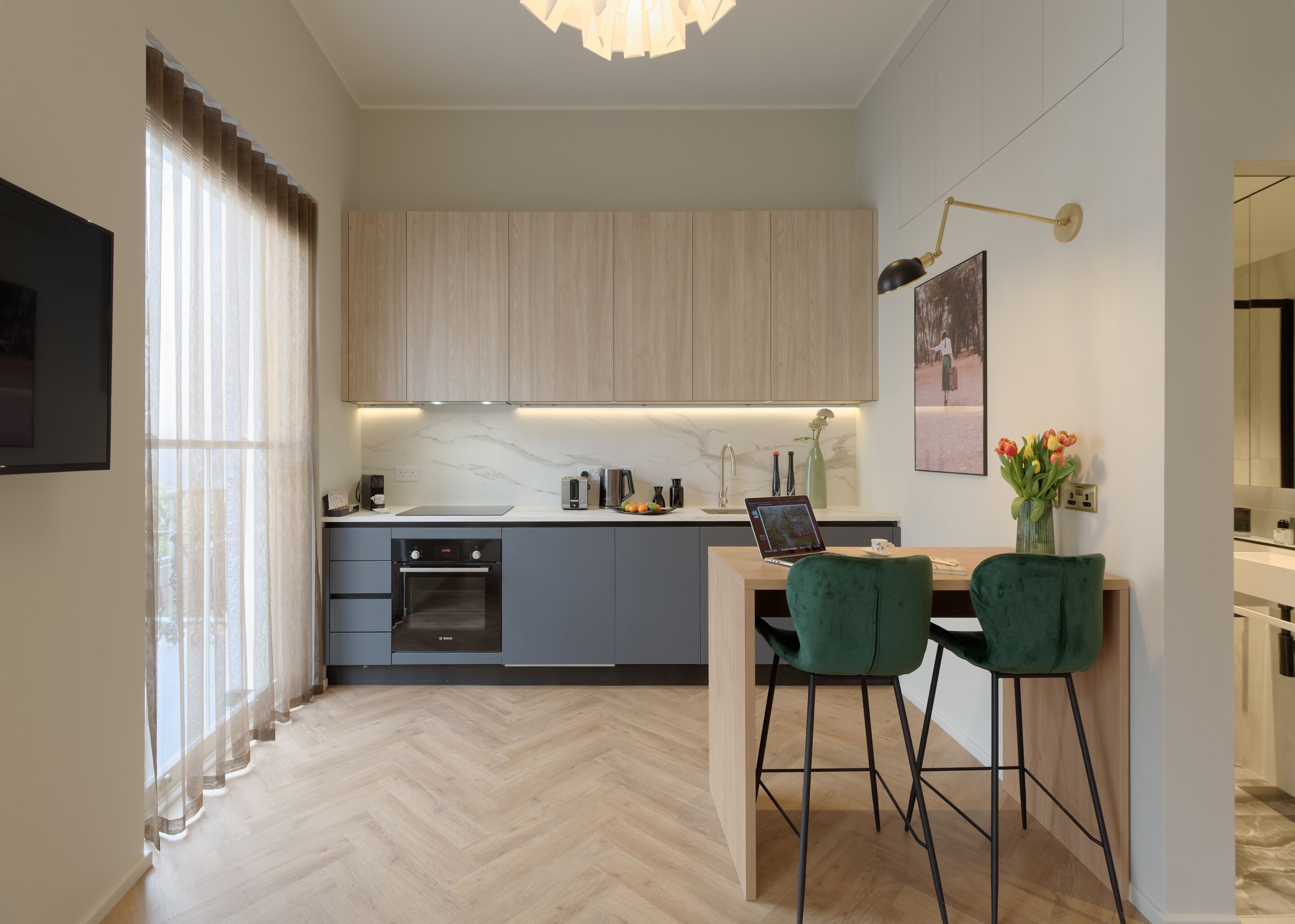 Une cuisine moderne dotée d’un four encastré, d’un coin repas avec deux chaises vertes et d’un luminaire suspendu au-dessus du comptoir.