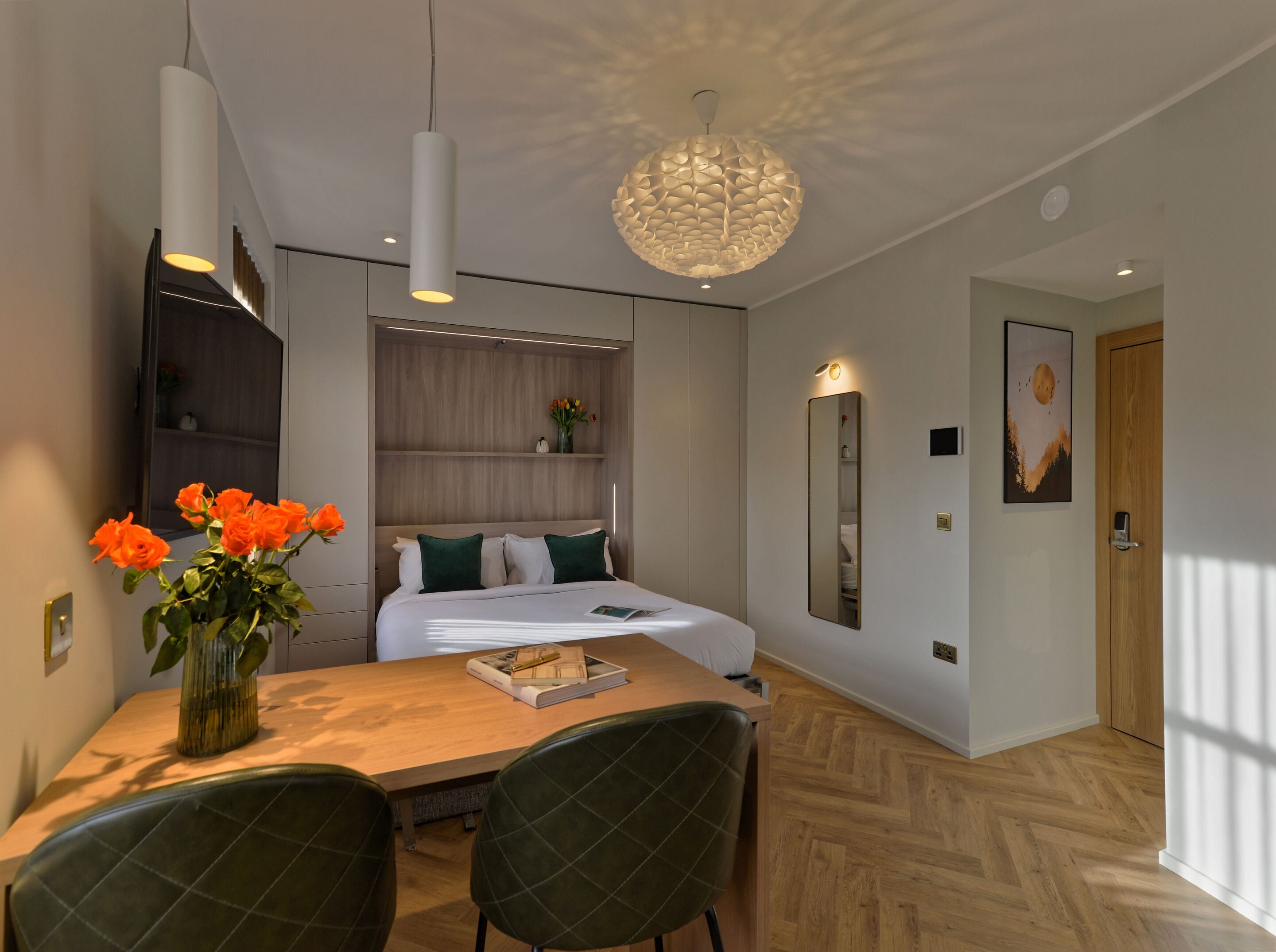 A modern hotel room with a large bed, a dining table with chairs, a vase of flowers, and a large mirror on the wall.
