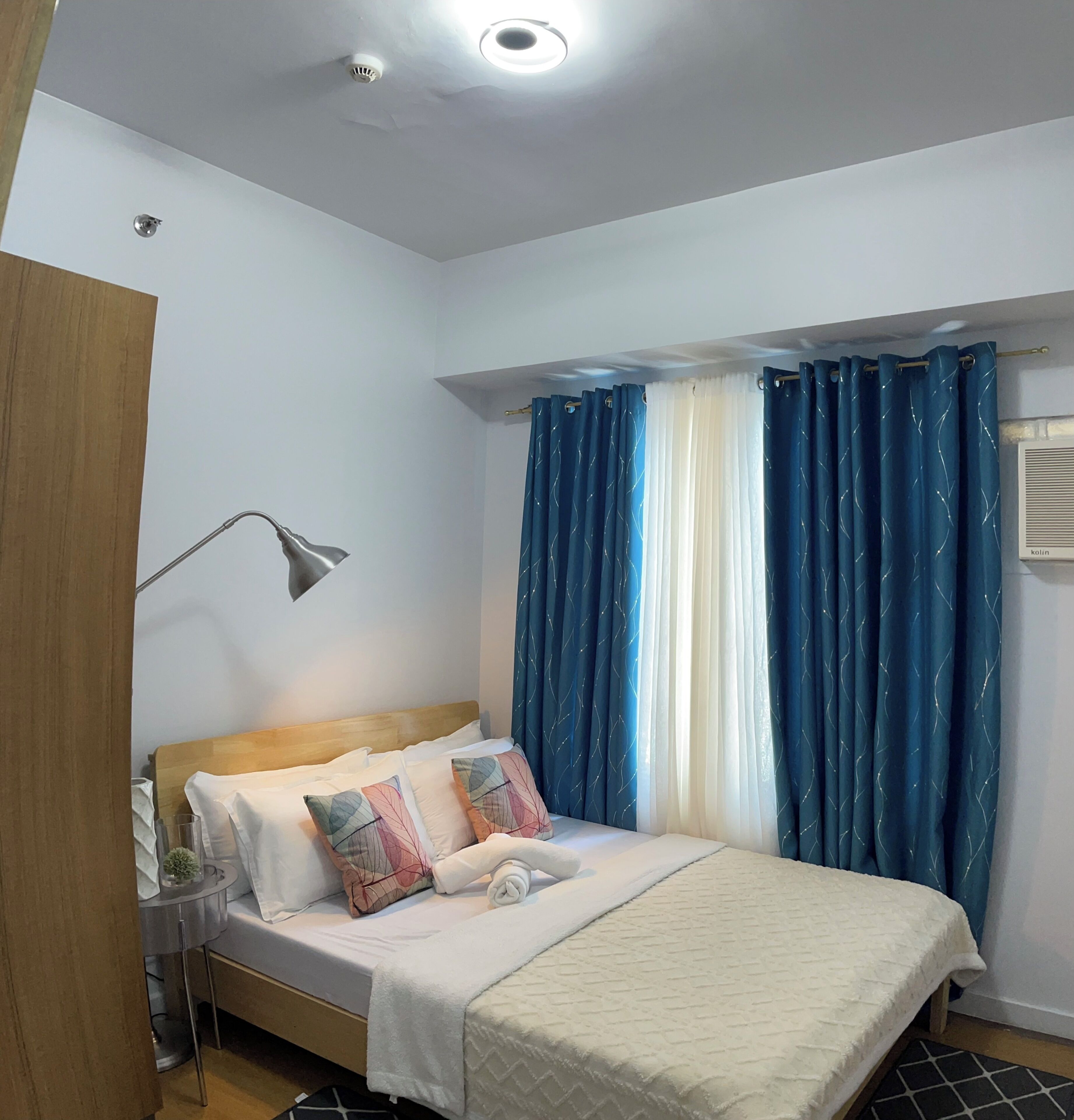 Una cama bien hecha con almohadas, una lámpara de noche y una ventana con cortinas azules.