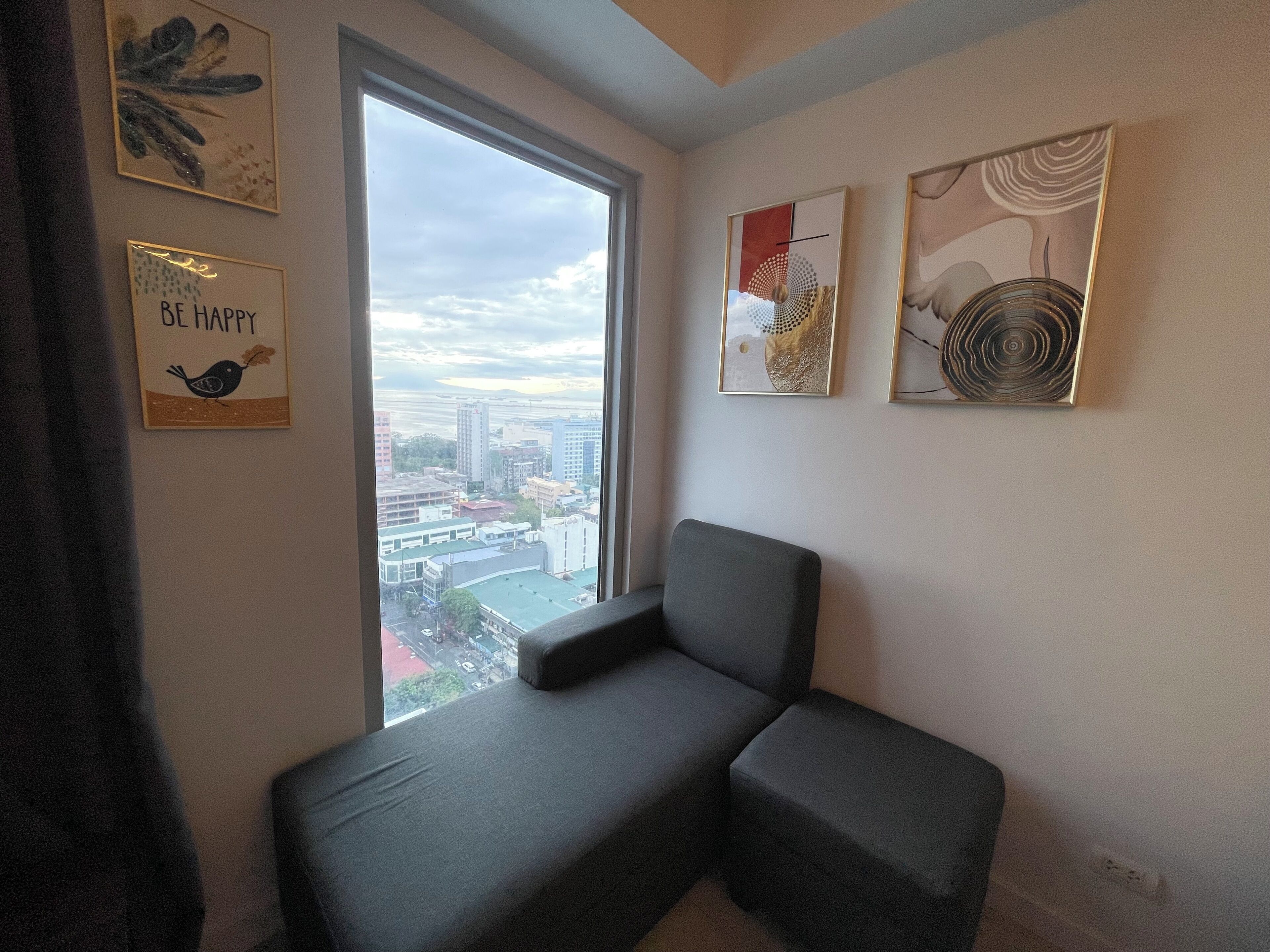 A corner room with a grey sofa, a large window with a city view, and three framed artworks on the wall.