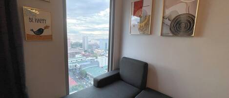 A corner room with a grey sofa, a large window with a city view, and three framed artworks on the wall.