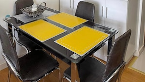 A compact dining area with a glass table, yellow placemats, and black chairs. Above the table, there is a white cabinet with open shelves and a mirror. A painting of a coastal town is on the wall to the left. - Modern Studio with Sunset View  (Manila)