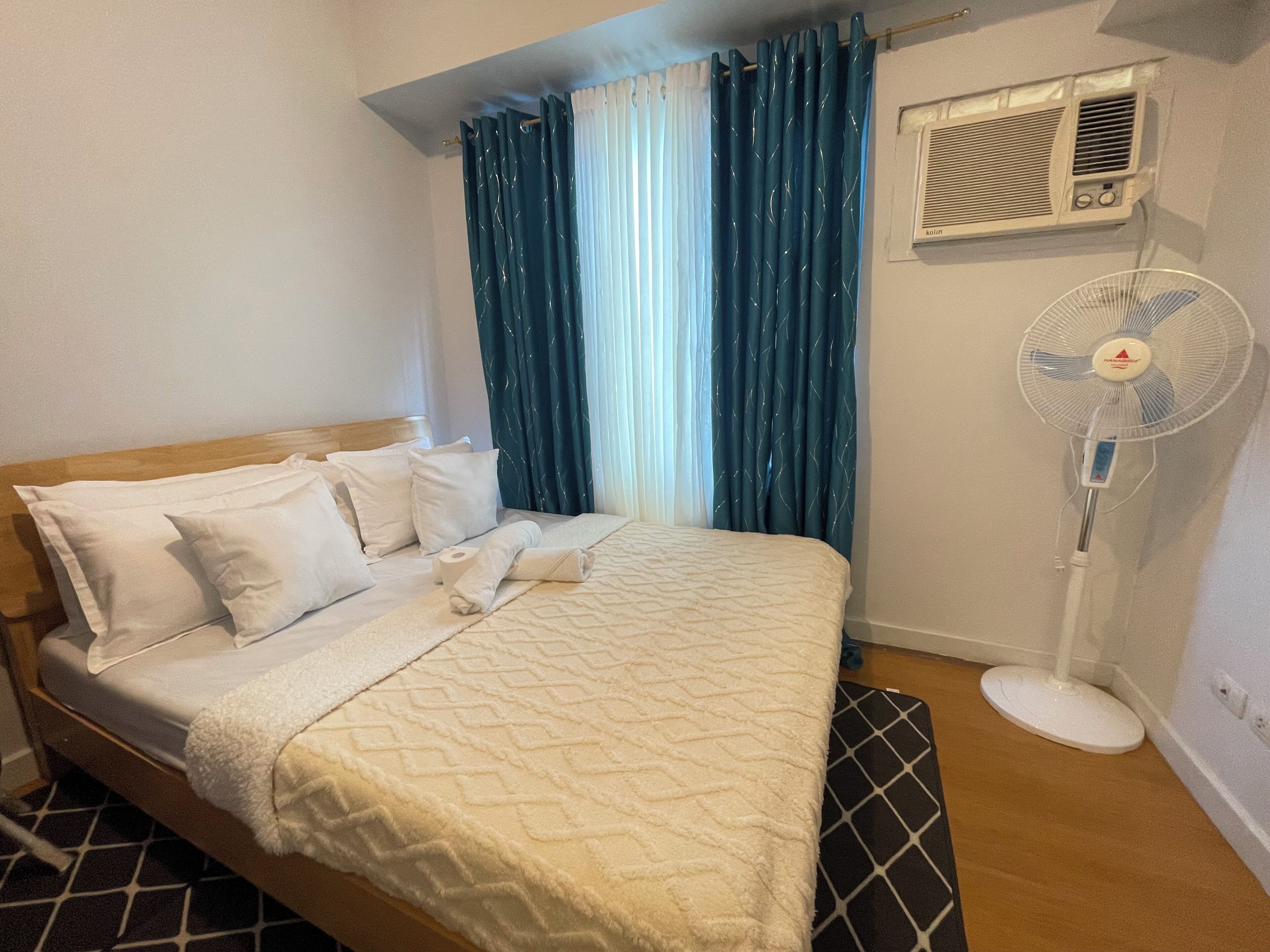 A hotel room with a bed, white pillows, a rolled towel, a standing fan, and a window with blue curtains.