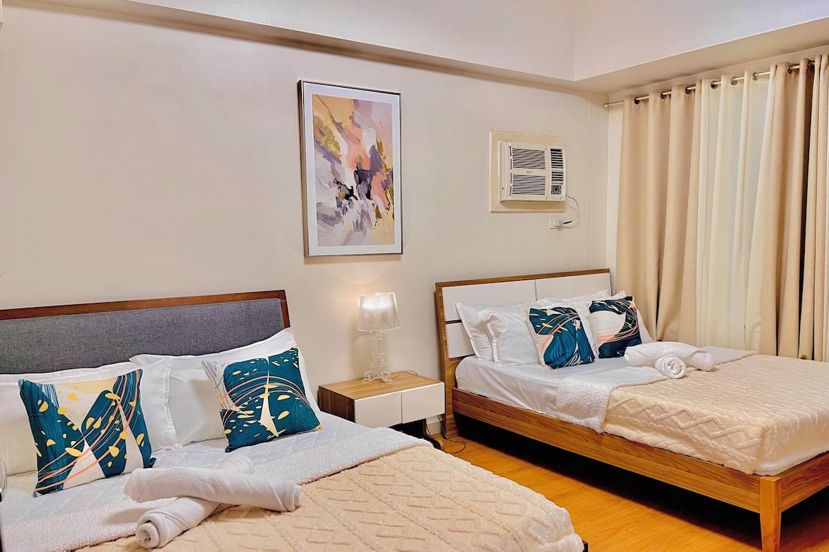 Two beds with headboards, a bedside table with a lamp, a wall-mounted air conditioner, and a window with curtains.
