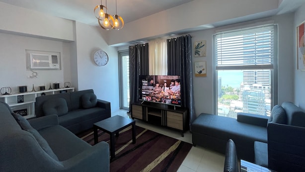 A modern living room with a flat-screen TV, sofas, a coffee table, and a large window with blinds. - Modern Studio with Sunset View  (Manila)