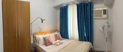 A bedroom with a bed, a nightstand, a lamp, a wardrobe, a window with curtains, an air conditioner, and a standing fan.