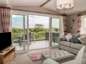 A modern living room with a large sliding glass door leading to a balcony, a grey sofa with cushions, a pink rug, and a television on a wooden stand. - Maple Lodge (Newton Abbot)