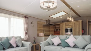 A modern living room with two sofas, a small table, and a large ottoman. A kitchen is visible in the background with wooden cabinets and a built-in oven. - Maple Lodge (Newton Abbot)