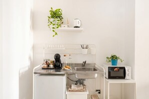 Une petite cuisine équipée d’un évier, d’une machine à café, d’un four à micro-ondes et d’une petite étagère sur laquelle sont posés une plante et une bouilloire.