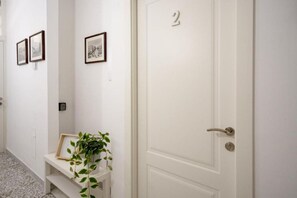 Un couloir avec une porte blanche portant le numéro 2, un banc avec un pot de fleurs et des tableaux encadrés accrochés aux murs.