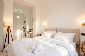 A neatly made bed with white linens and rolled towels, a bedside table with a lamp, and a view into a dining area.