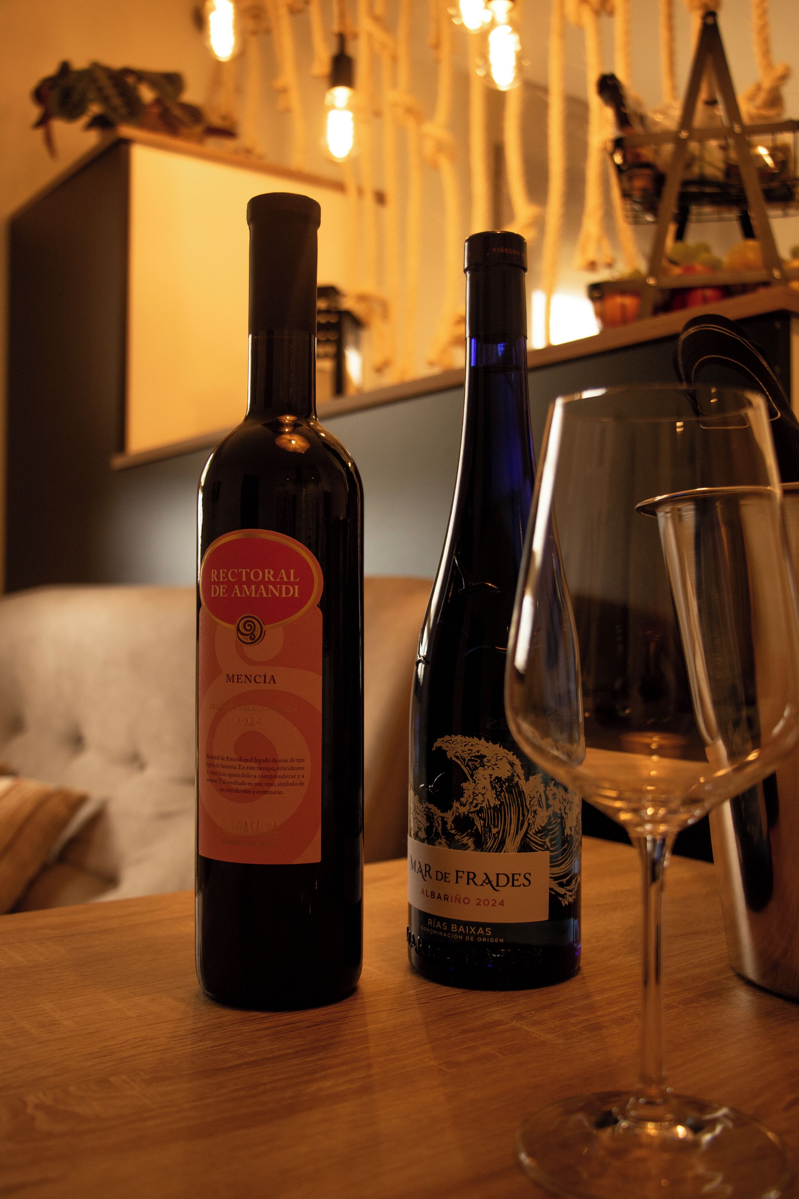 Dos botellas de vino y una copa de vino vacía sobre una mesa de madera en una habitación con luz cálida.
