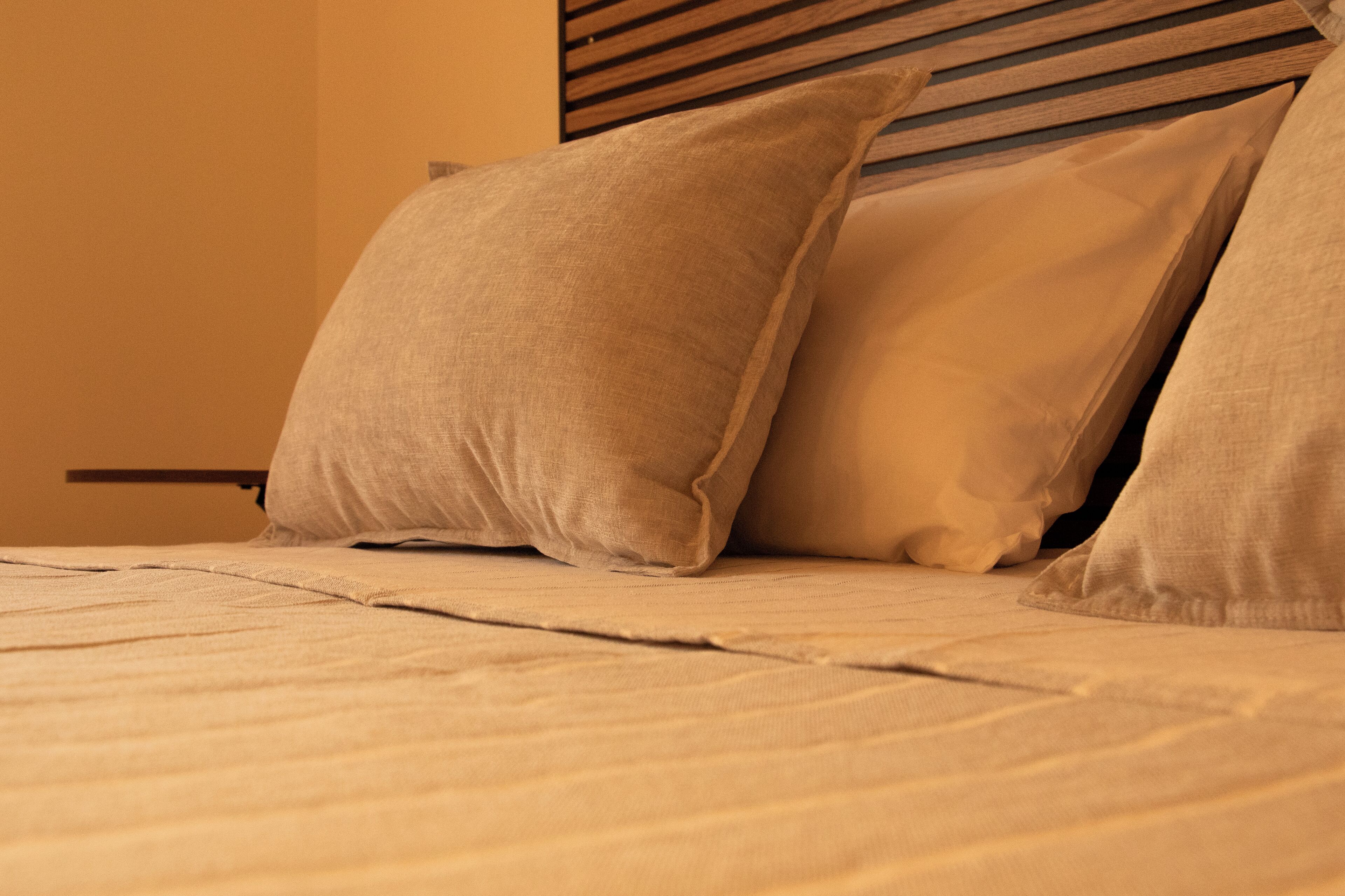 A bed with beige bedding and pillows, a wooden headboard, and a plain wall.