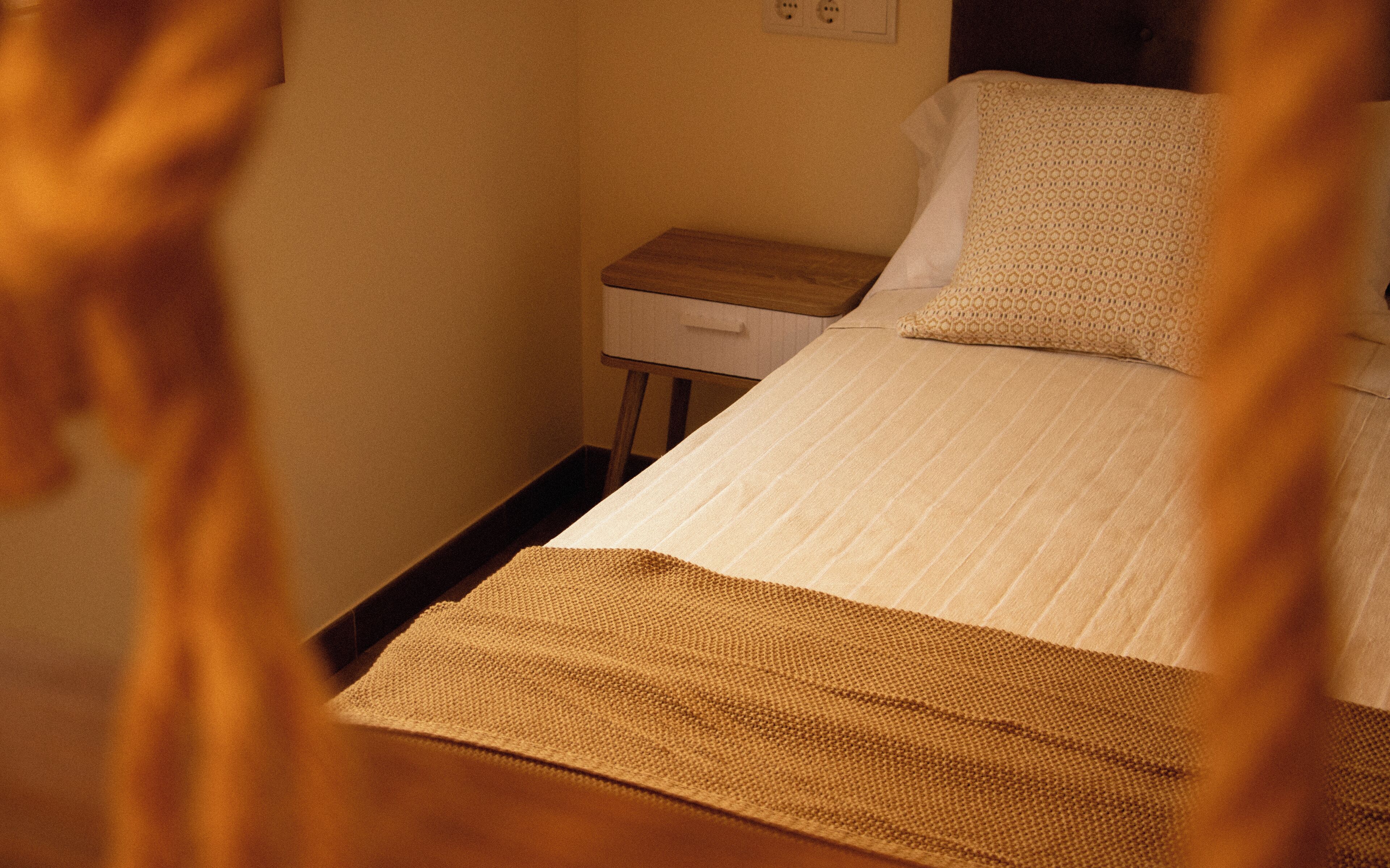 A small, neatly made bed with a white sheet and a brown blanket, a wooden nightstand with a drawer, and a wall socket.