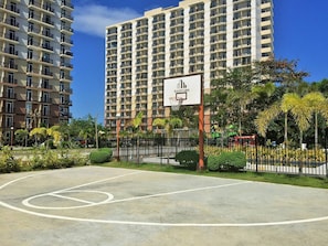 A basketball court with a sign, surrounded by a fence and greenery, with high-rise buildings in the background.