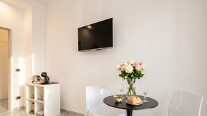 A modern room with a flat-screen TV, a round table with two chairs, a small white cabinet, and a vase with flowers.