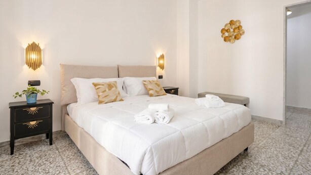 A neatly made bed with white bedding, two bedside tables with wall-mounted lamps, a decorative wall piece, and a potted plant on one of the nightstands.