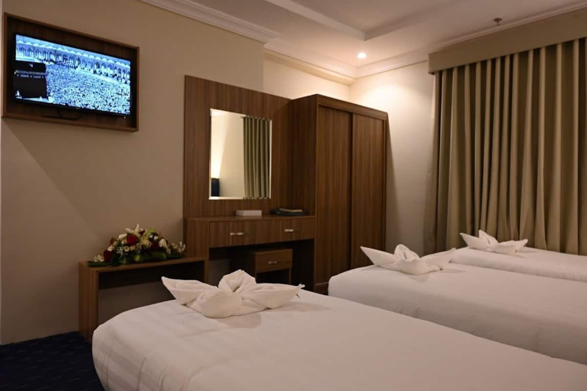 A hotel room with two beds, a TV mounted on the wall, a wooden desk with a mirror, and a window with curtains.