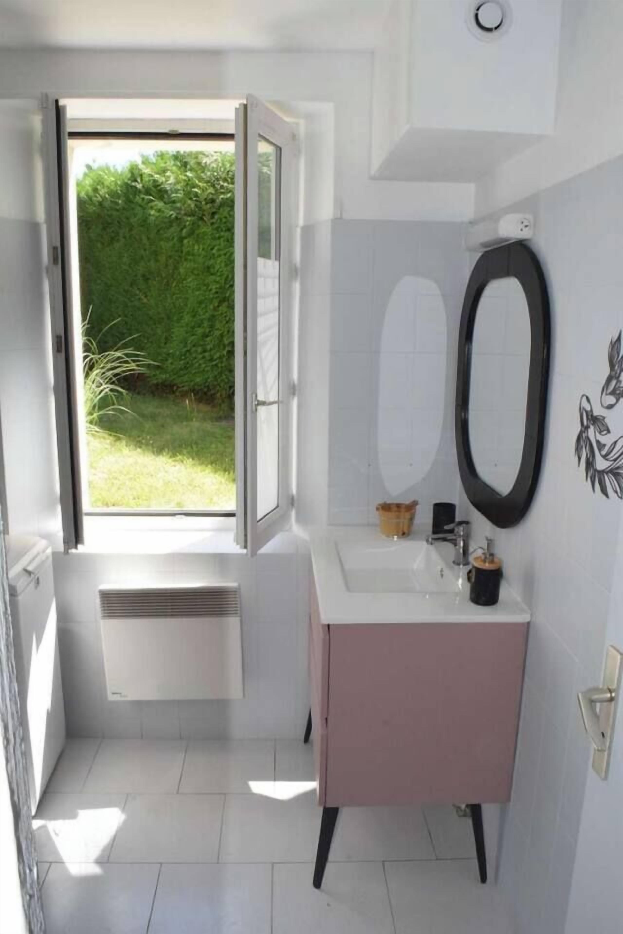 Ein Badezimmer mit einem weißen Waschbecken, einem Spiegel und einem Fenster mit Blick auf Grünfläche.