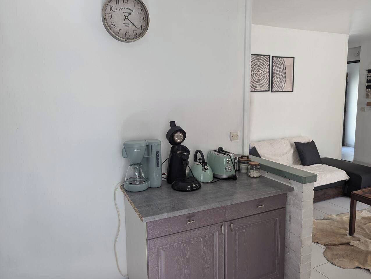 Una cocina con cafetera, hervidor, tostadora y hervidor sobre una encimera. Hay un reloj en la pared y un sofá al fondo.