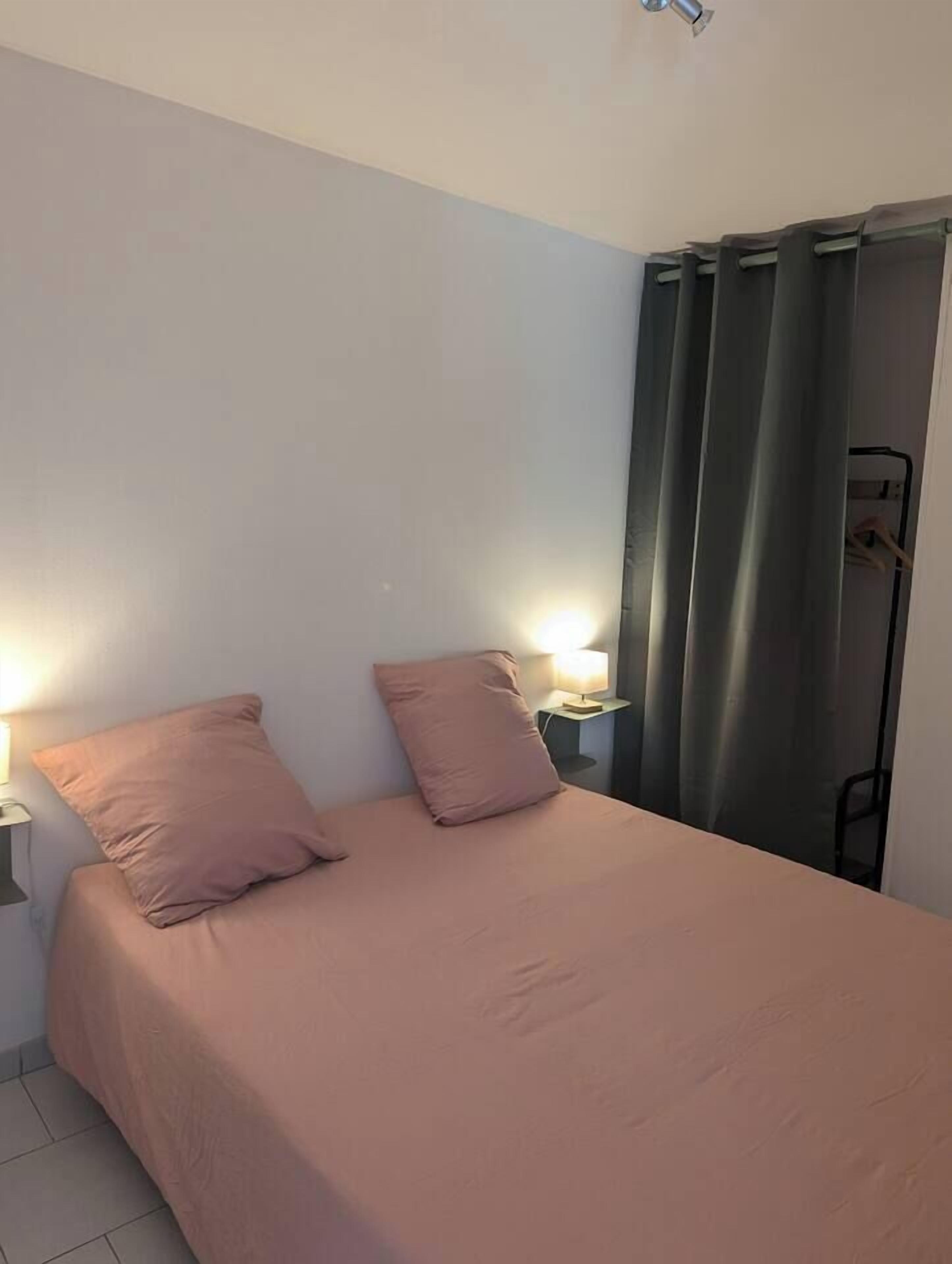 Een eenpersoonskamer met een roze sprei, twee kussens, een nachtkastje met een lamp en een gordijn voor het raam.