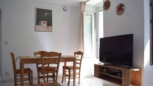 A living room with a wooden dining table, four chairs, a TV on a wooden stand, a painting on the wall, and a window with curtains. - The Bangor Coon Apartment Liouba (Forges)
