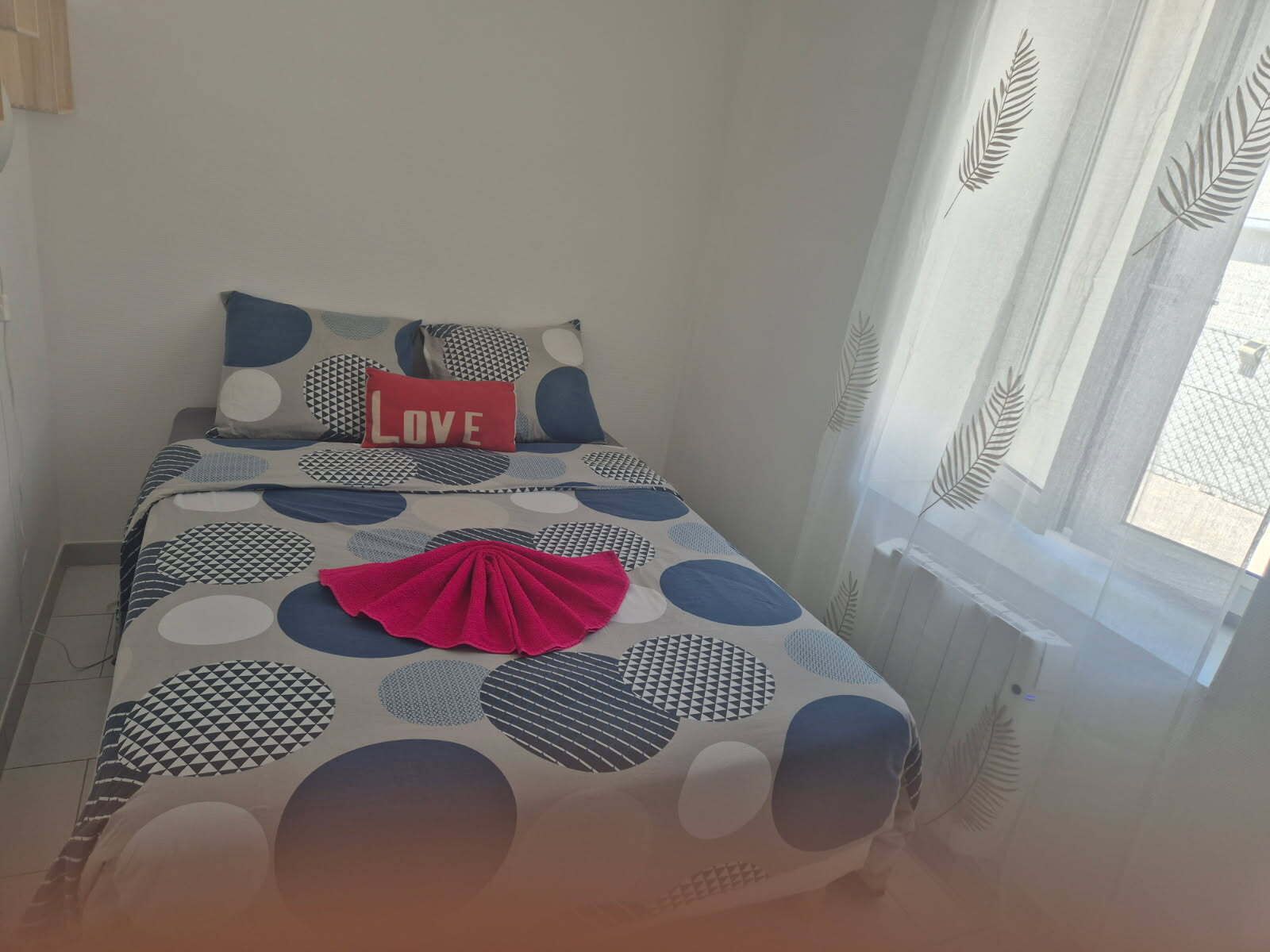 Ein kleines, ordentlich gemachtes Bett mit einer geometrisch gemusterten Tagesdecke, einem roten Kissen mit dem Schriftzug "LOVE" und einem Fenster mit transparenten Vorhängen.