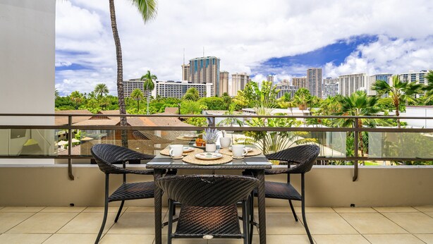 A balcony with a table set for two, overlooking a cityscape with palm trees and high-rise buildings. - Heart of Waikiki, 2 blocks to beach, kitchenette, lanai, free parking nearby! (Honolulu)