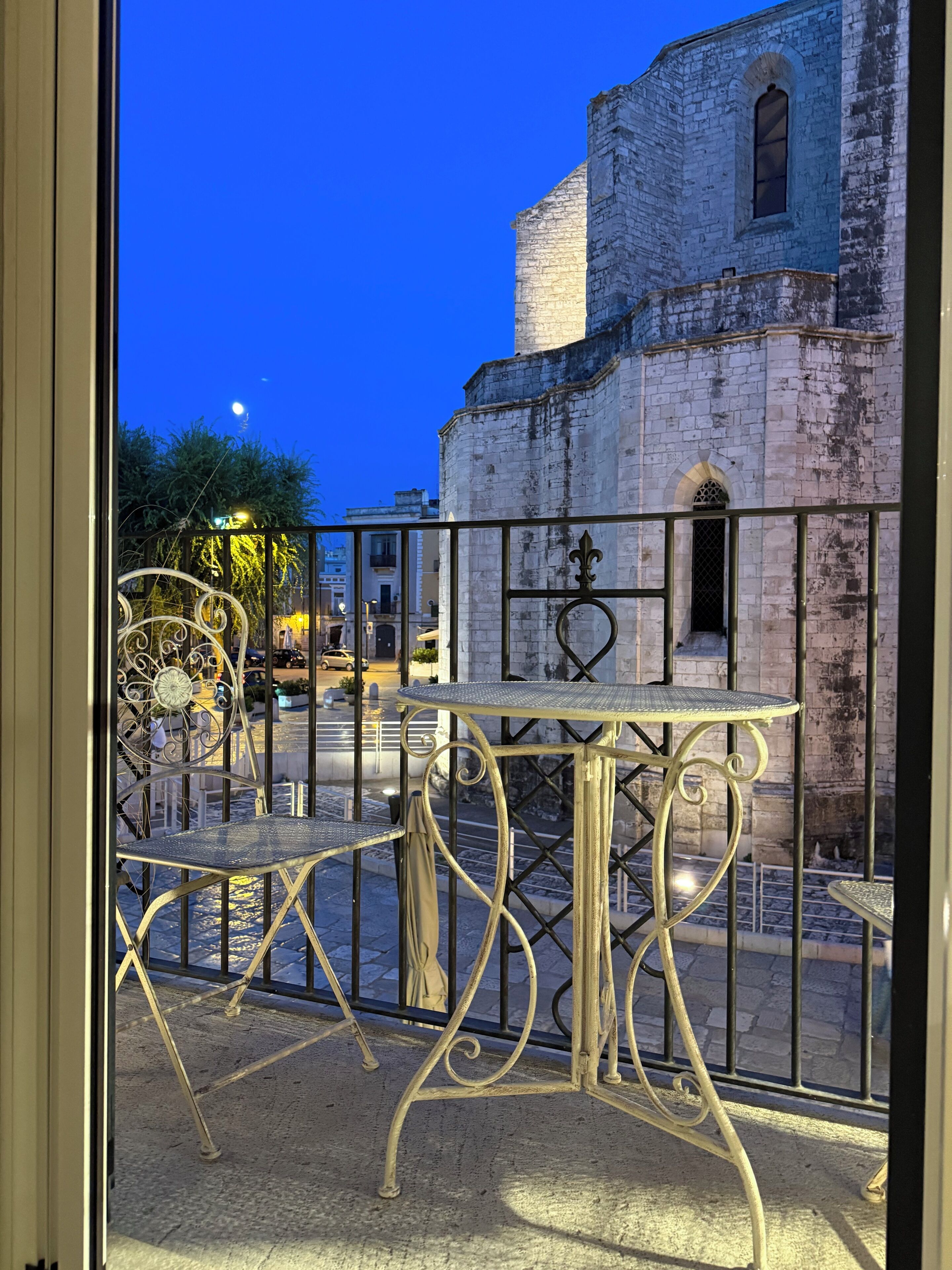 A balcony with wrought iron furniture, a stone building with a clock tower in the background, and a clear night sky.