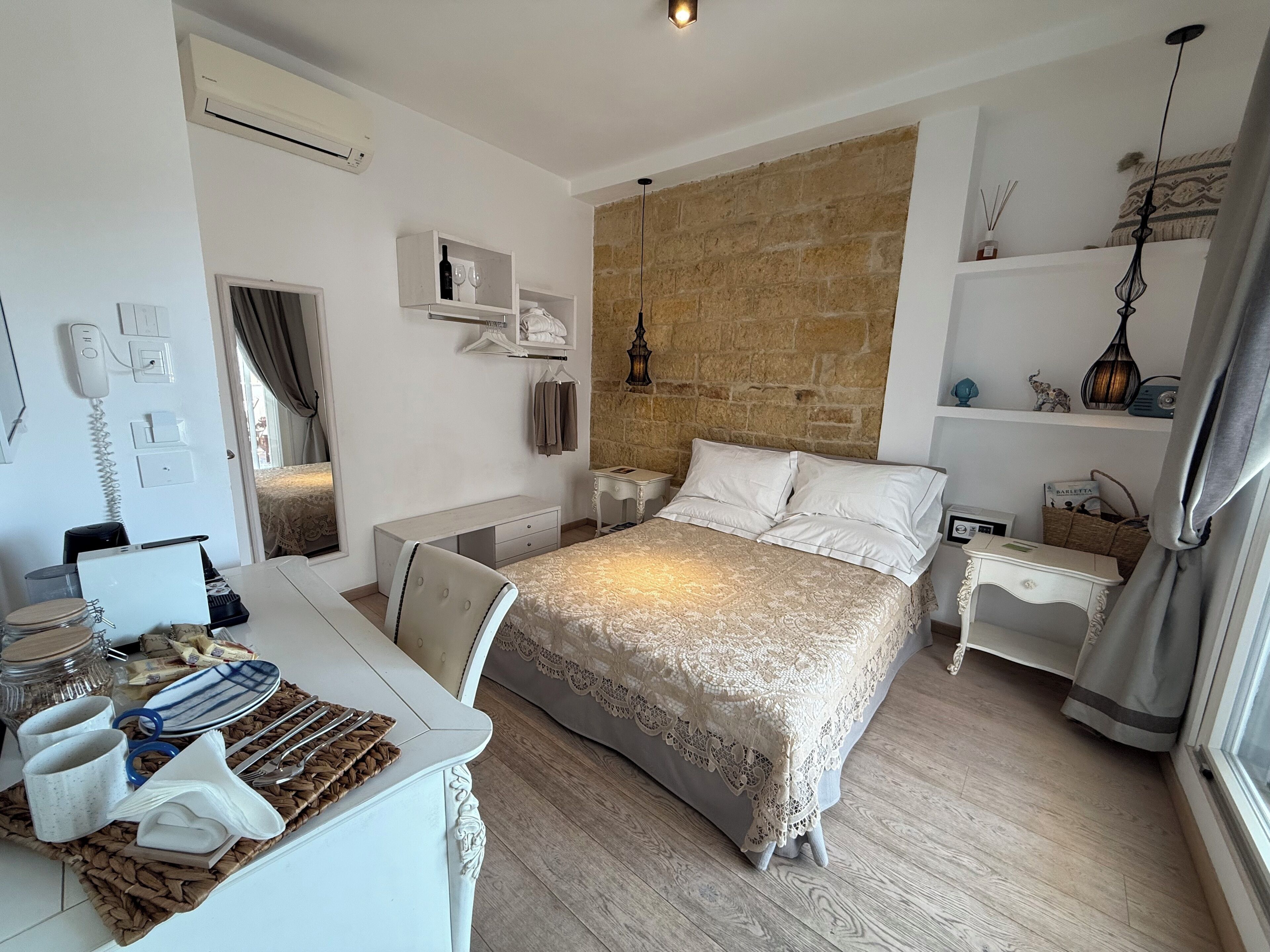 A bedroom with a bed, bedside tables, a desk with a chair, a wardrobe, and a stone wall.