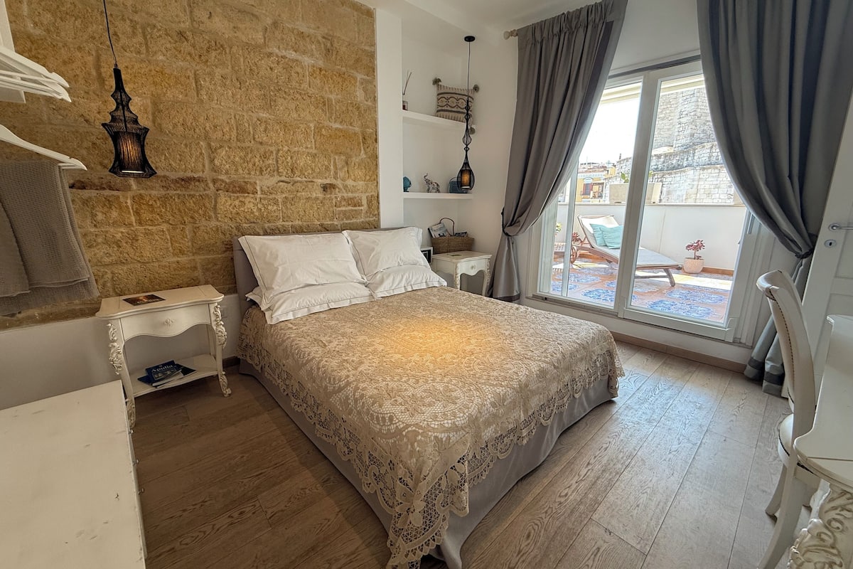 Ein Schlafzimmer mit einem großen Bett, Holzboden, Steinwand, einem Fenster mit Vorhängen und einem kleinen Nachttisch.
