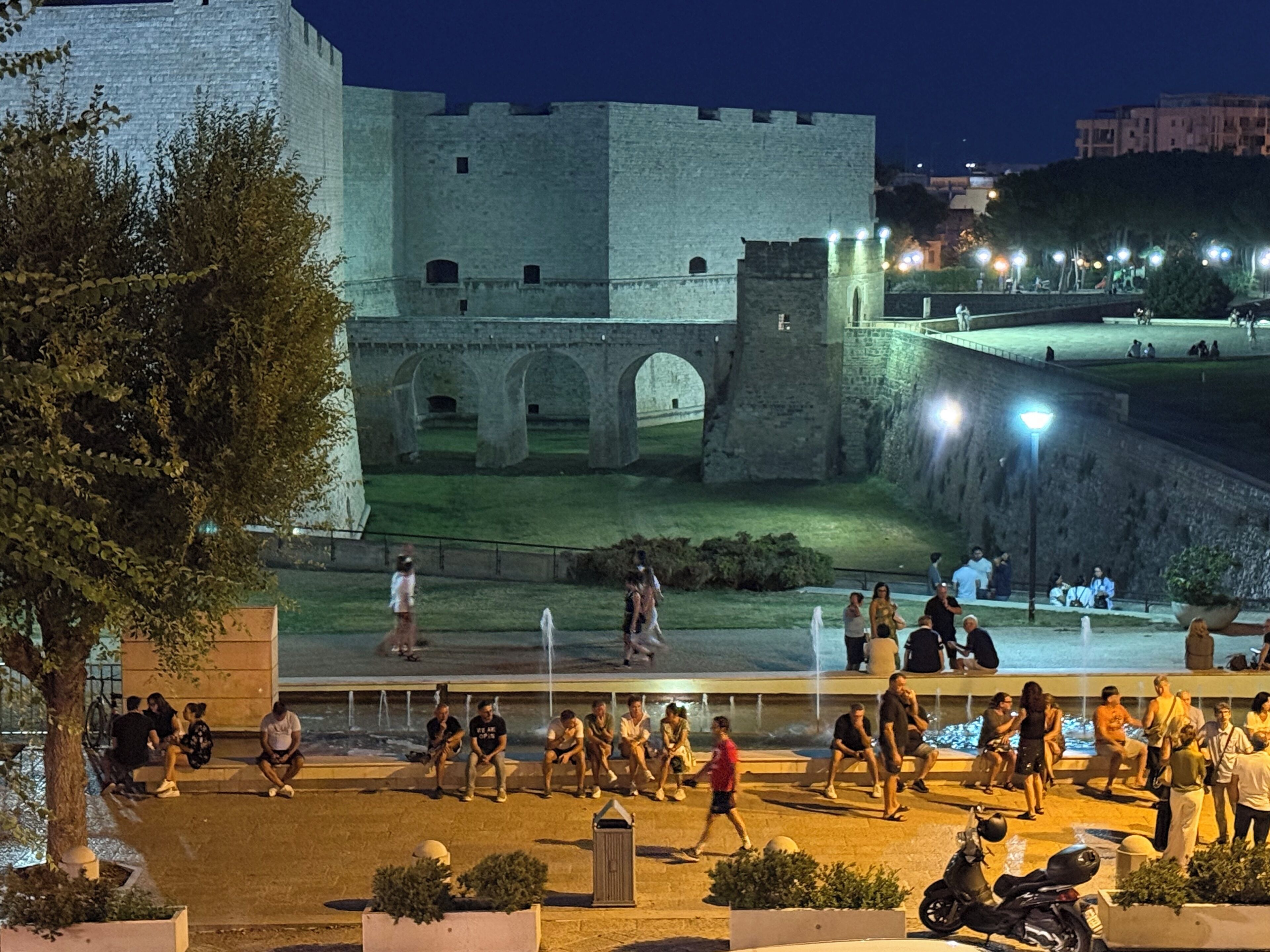 Un edificio histórico de piedra con arcos y un foso, rodeado por una plaza pública con gente y una fuente.