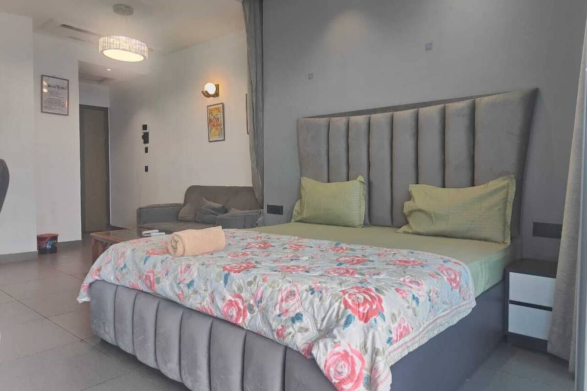 A modern bedroom with a large bed, grey upholstered headboard, and a floral bedspread. There is a bedside table with a lamp and a nightstand with a drawer.