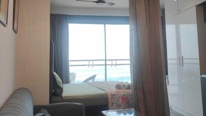A modern hotel room with a sofa, a glass-top coffee table, a bed with a floral bedspread, a ceiling fan, and a large window with curtains. - Supernova Heights Studio (Noida)